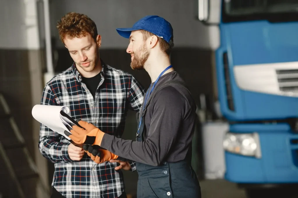 Technician in uniform consults with customer in garage setting beside a blue truck.